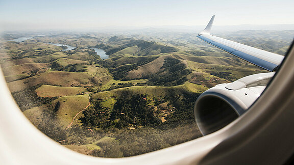 Blick auf eine grüne, leicht hügelige Landschaft aus dem Fenster einer Embraer E195 Blick auf eine grüne, leicht hügelige Landschaft aus dem Fenster einer Embraer E195